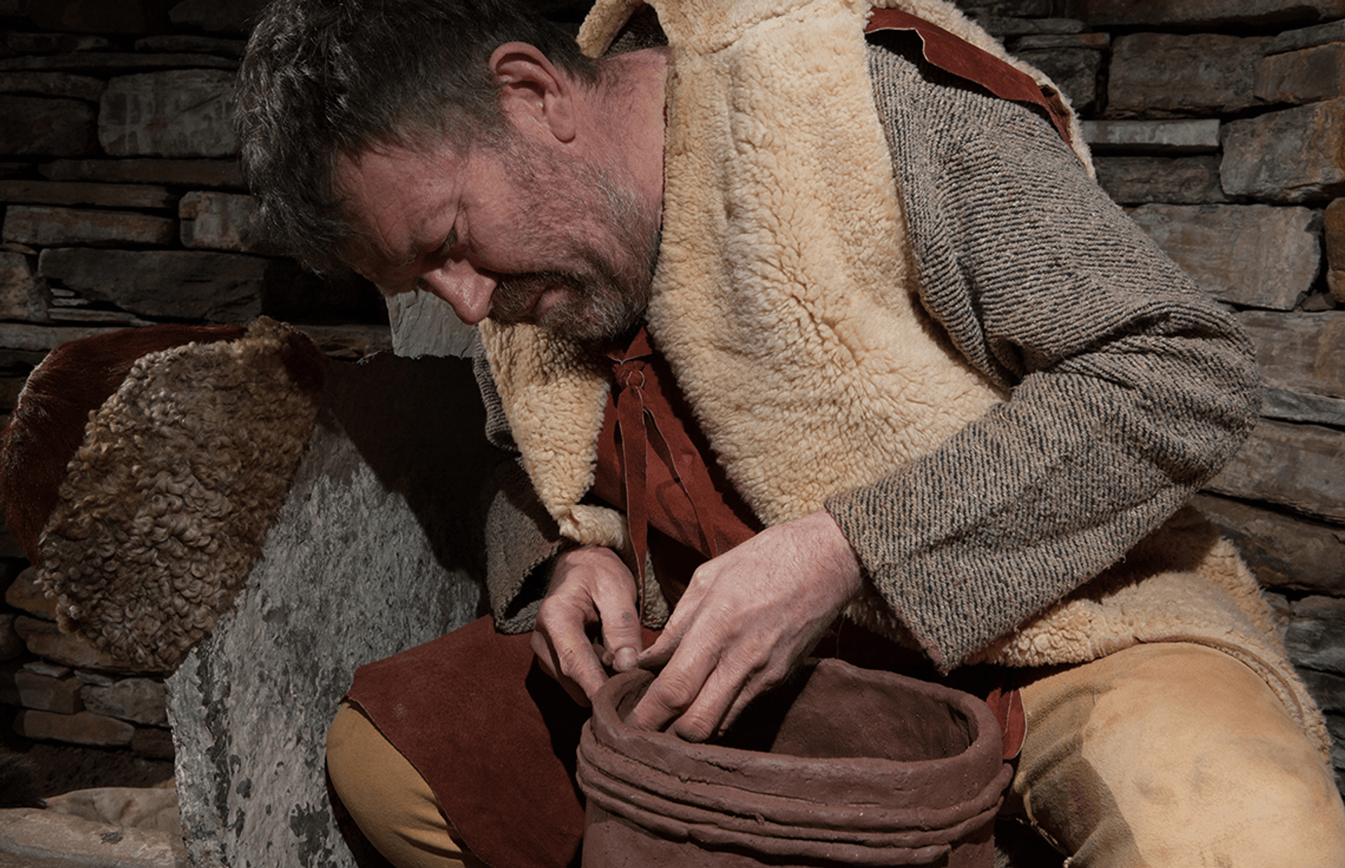 © Ken Amer
An older man with short grey hair, wearing cloth and animal skin clothes holds an unbaked clay pot as he decorates the rim. © Ken Amer
An older man with short grey hair, wearing cloth and animal skin clothes holds an unbaked clay pot as he decorates the rim.