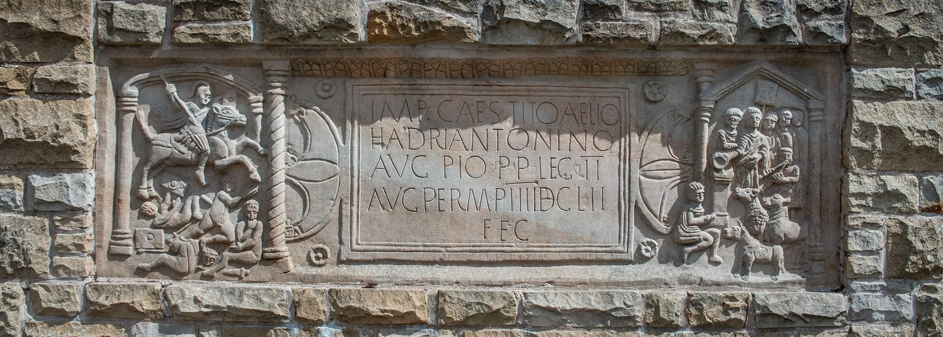 © Rediscovering the Antonine Wall Project
A sandstone distance stone built into a brick wall. The distance stone has a Latin inscription in its center, flanked on either side by carved images. © Rediscovering the Antonine Wall Project
A sandstone distance stone built into a brick wall. The distance stone has a Latin inscription in its center, flanked on either side by carved images.