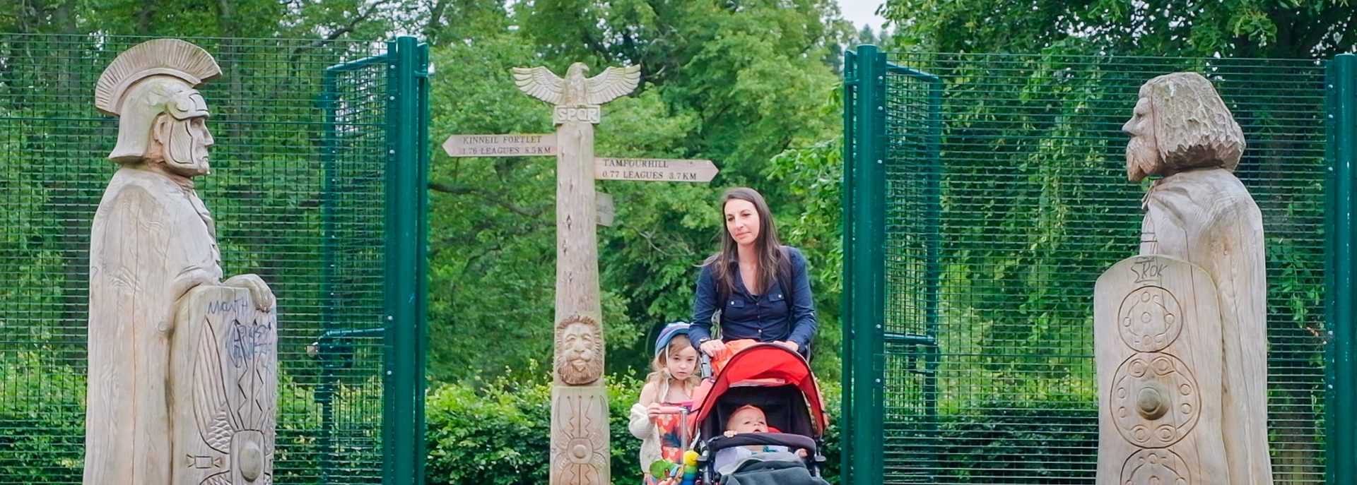 © Rediscovering the Antonine Wall Project.
A woman pushing a pram and accompanied by a child on a scooter enters through a green fenced gate. There is a wooden sculpture on either side of the group. © Rediscovering the Antonine Wall Project.
A woman pushing a pram and accompanied by a child on a scooter enters through a green fenced gate. There is a wooden sculpture on either side of the group.