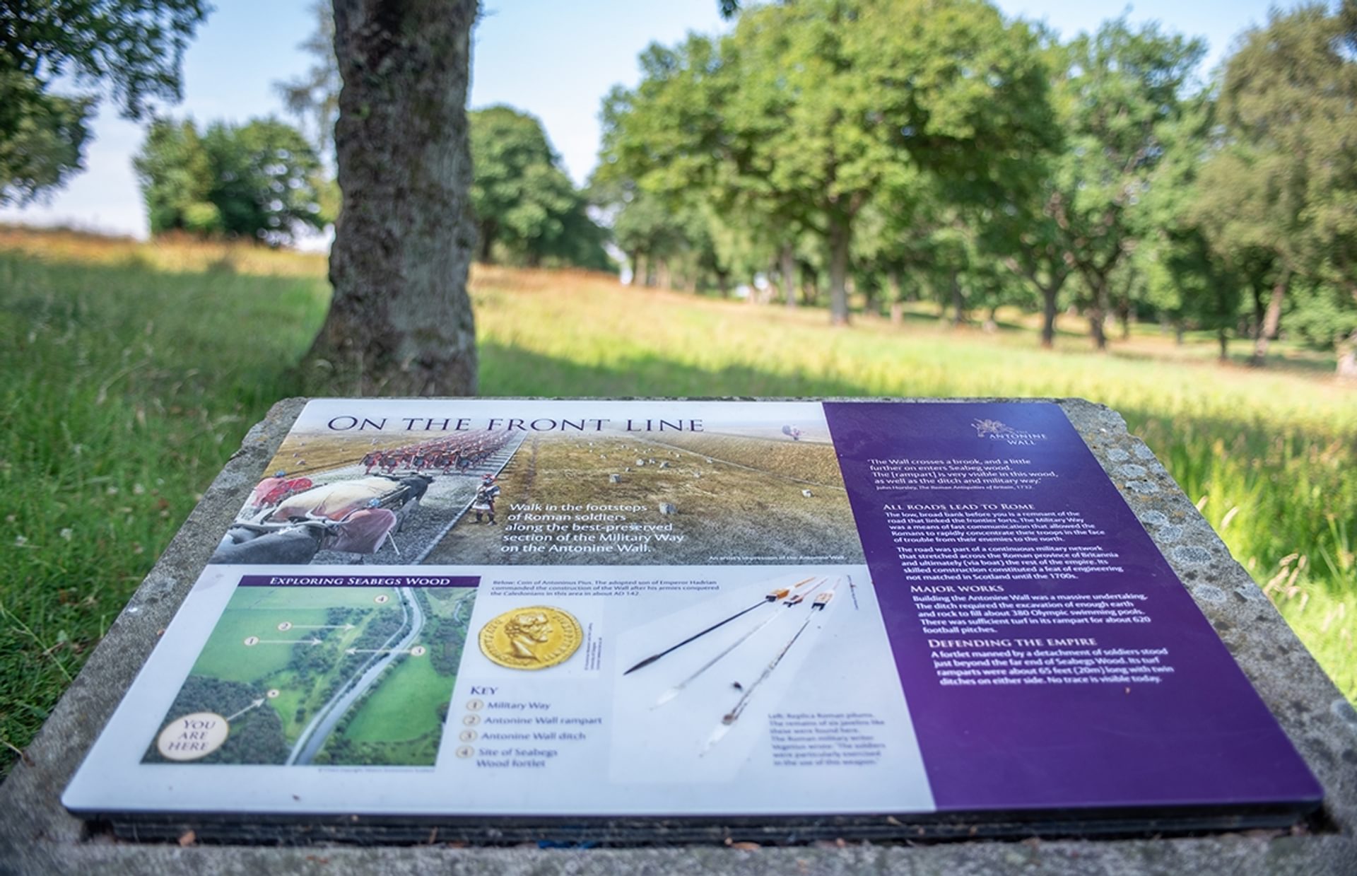 © Rediscovering the Antonine Wall Project
Seabegs Wood, a white and purple interpretation panel on a low stone plinth with grass, trees, and sky in the background. © Rediscovering the Antonine Wall Project
Seabegs Wood, a white and purple interpretation panel on a low stone plinth with grass, trees, and sky in the background.