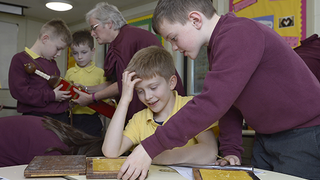 © Neil Hanna.
Two children at a classroom table using Roman replica items. A group of people in the background look at a Roman sword. © Neil Hanna.
Two children at a classroom table using Roman replica items. A group of people in the background look at a Roman sword.