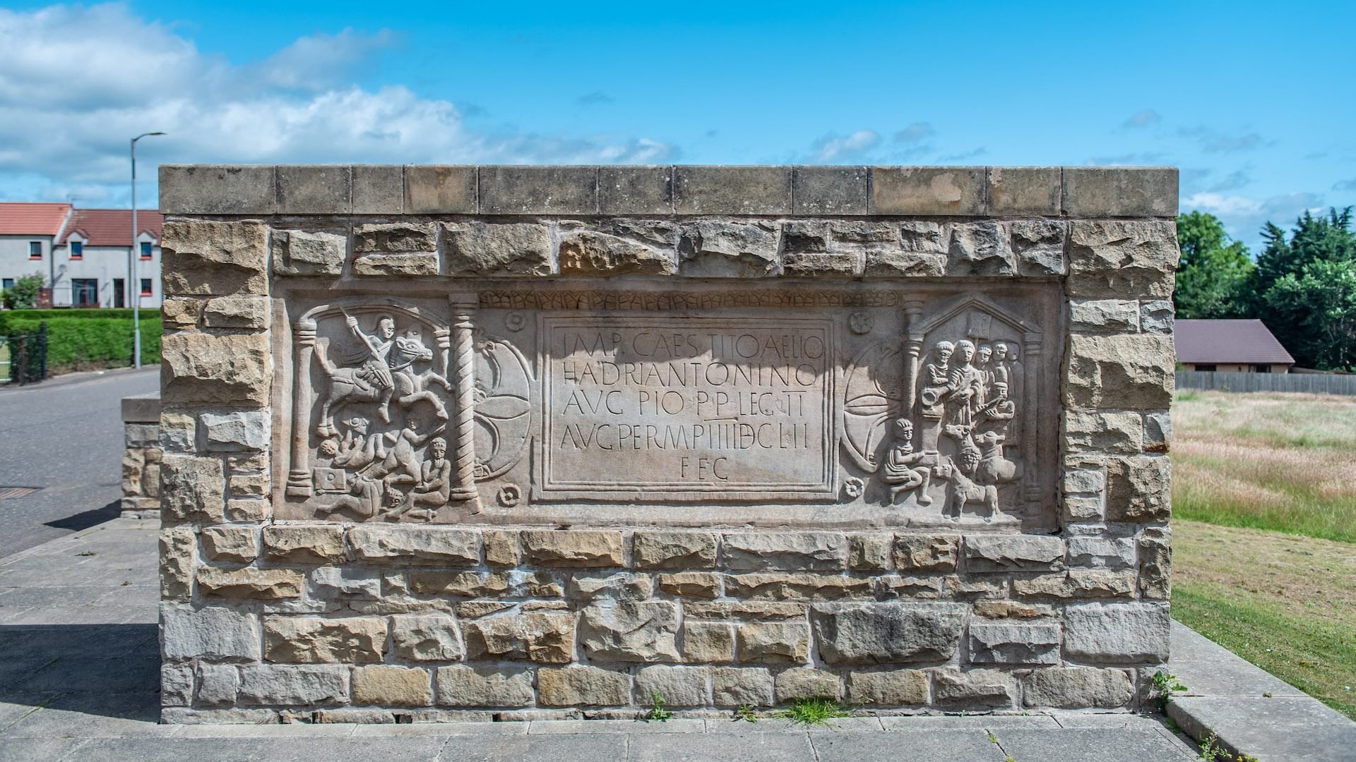 © Rediscovering the Antonine Wall
The replica Bridgeness Slab. A rectangle of sandstone carved in high relief set in a stone wall. Grass and blue sky beyond. © Rediscovering the Antonine Wall
The replica Bridgeness Slab. A rectangle of sandstone carved in high relief set in a stone wall. Grass and blue sky beyond.