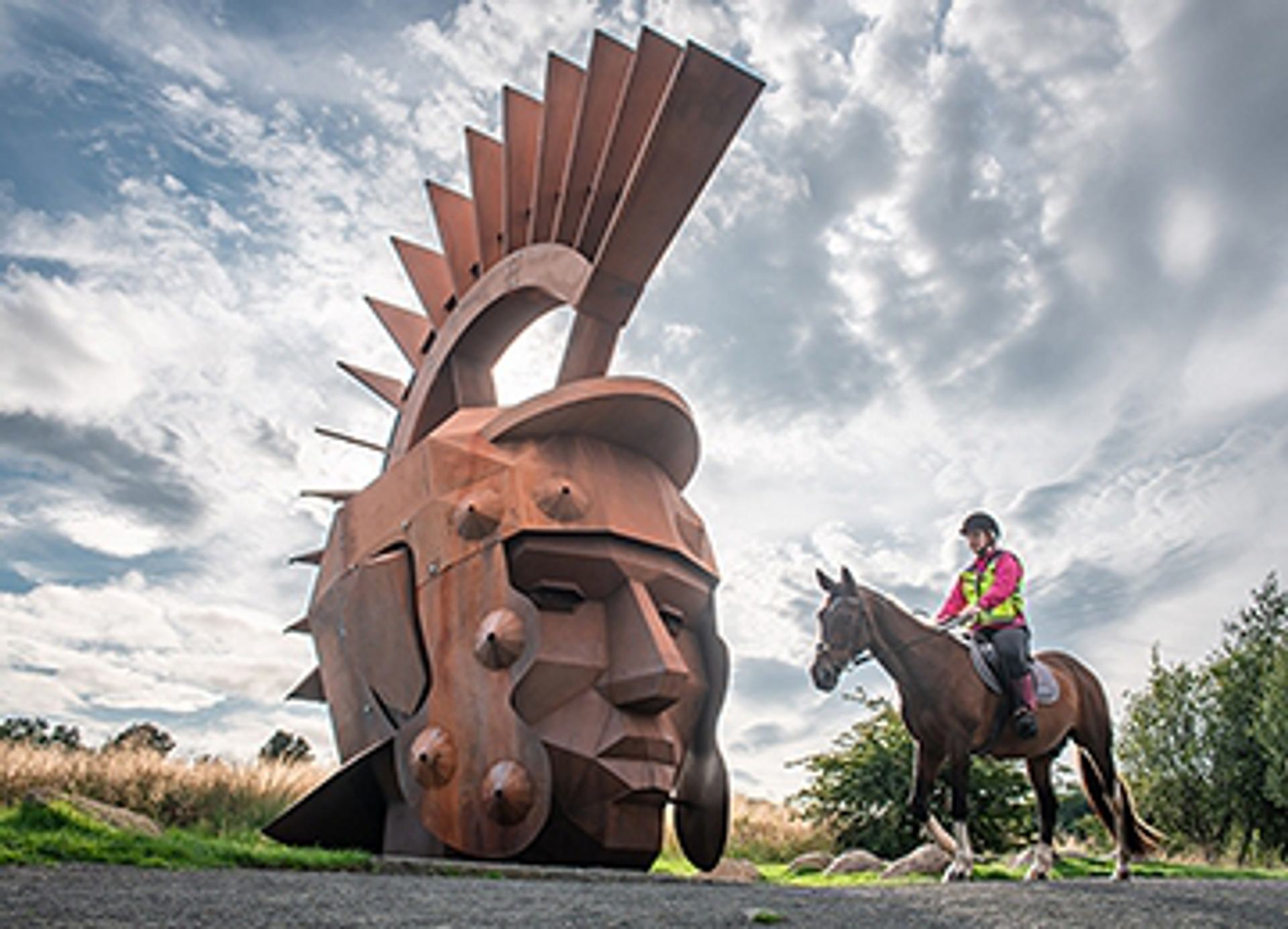 The Nethercroy Silvanus sculpture. A rusty steel legionary head on a hill with a cloudy sky. A horse and rider to the right. The Nethercroy Silvanus sculpture. A rusty steel legionary head on a hill with a cloudy sky. A horse and rider to the right.