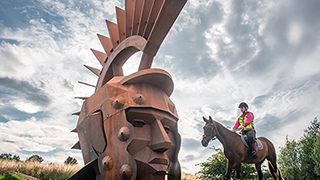 The Nethercroy Silvanus sculpture. A rusty steel legionary head on a hill with a cloudy sky. A horse and rider to the right. The Nethercroy Silvanus sculpture. A rusty steel legionary head on a hill with a cloudy sky. A horse and rider to the right.