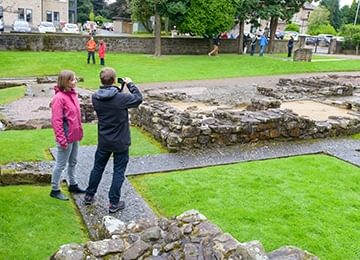 © Historic Environment Scotland
A couple take photos of the stone walls of Bearsden bathhouse. The walls are low and surrounded by lawn and a perimeter wall. © Historic Environment Scotland
A couple take photos of the stone walls of Bearsden bathhouse. The walls are low and surrounded by lawn and a perimeter wall.